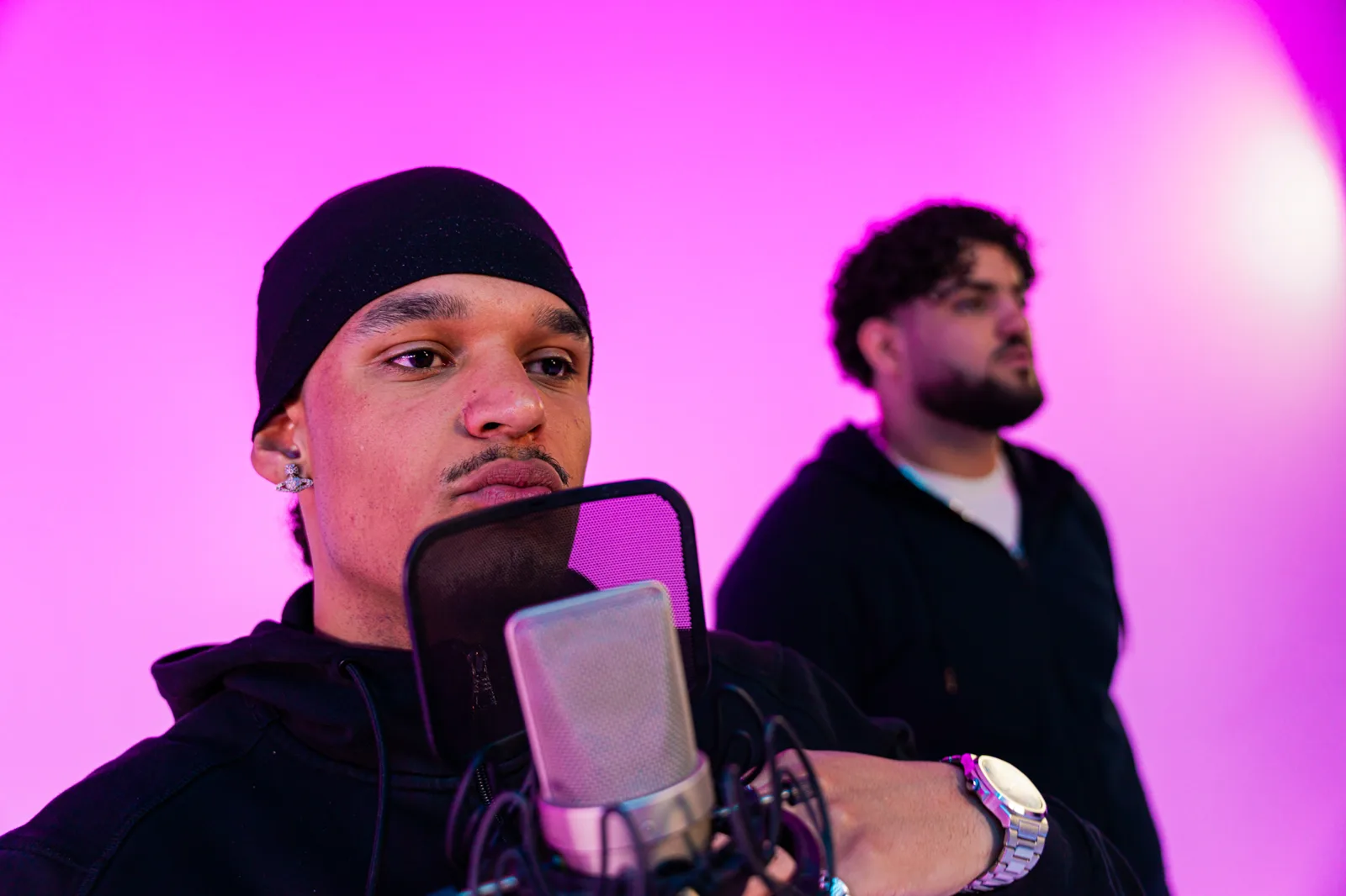Rapper met muts achter de microfoon, een tweede artiest op de achtergrond, roze achtergrond
